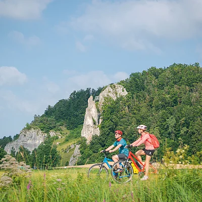 Zwei Personen fahren mit Fahrrädern auf einem Weg vor einer bewaldeten Felslandschaft.