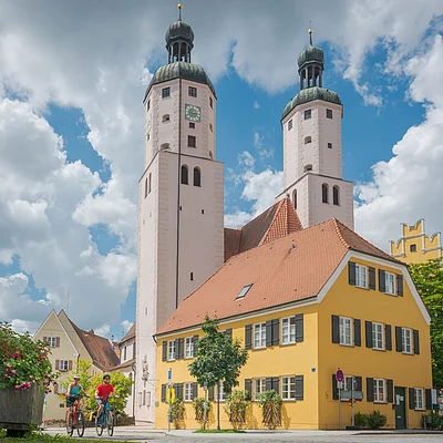 Zwei Radfahrer fahren vor einem gelben Gebäude mit roten Ziegeldächern und einer Kirche mit zwei Türmen im Hintergrund.