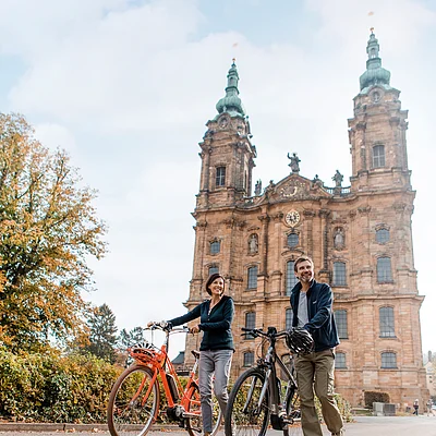 Zwei Personen mit Fahrrädern vor einer großen historischen Kirche an einem sonnigen Tag.