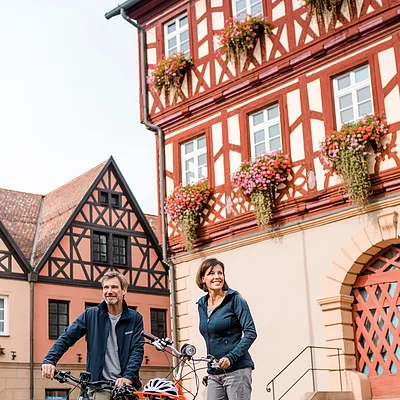 Mann und Frau mit Fahrrädern stehen auf gepflastertem Platz vor Fachwerkhäusern mit Blumenkästen.