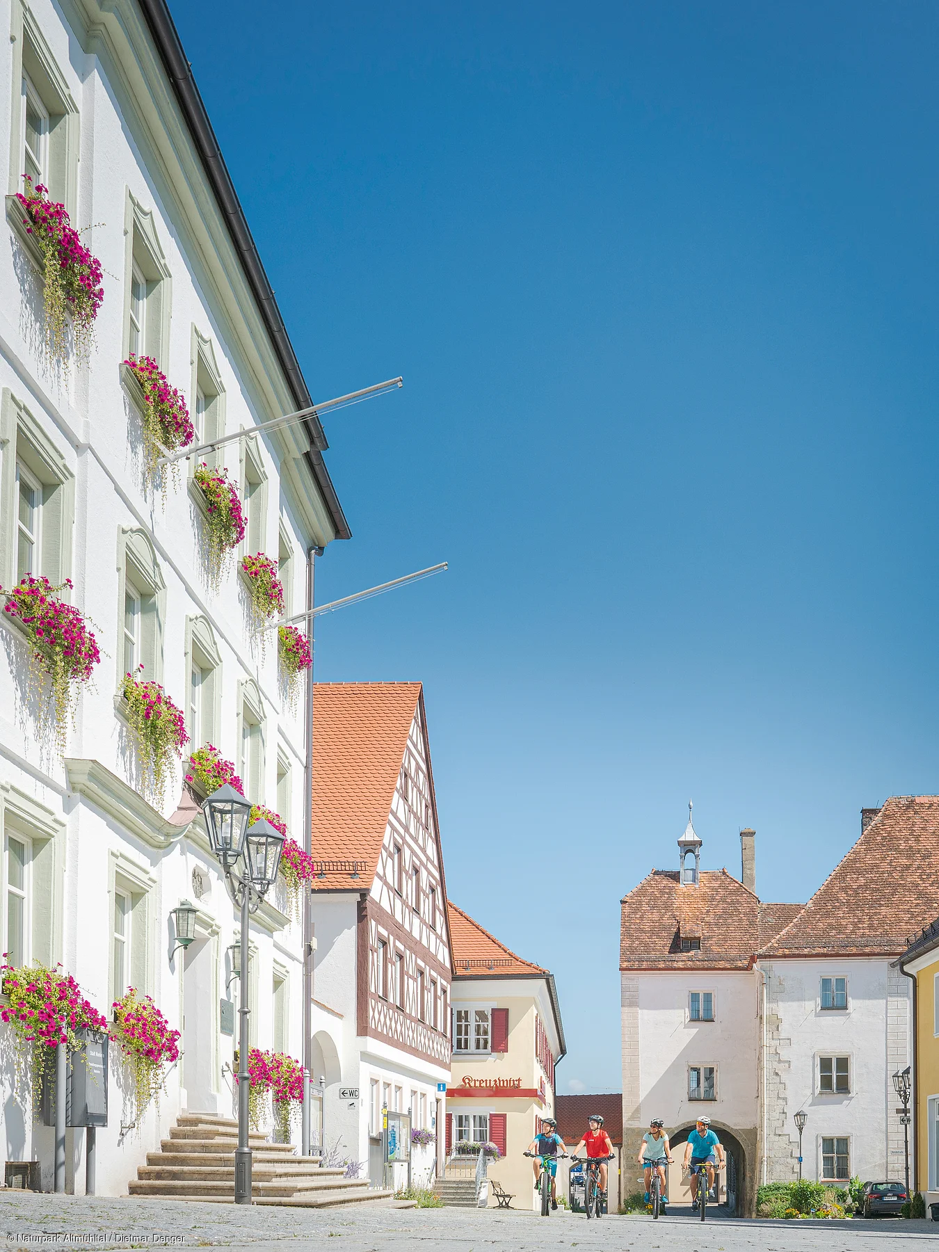 Historische Gebäude mit Blumenkästen und vier Radfahrern auf gepflastertem Platz bei klarem Himmel.