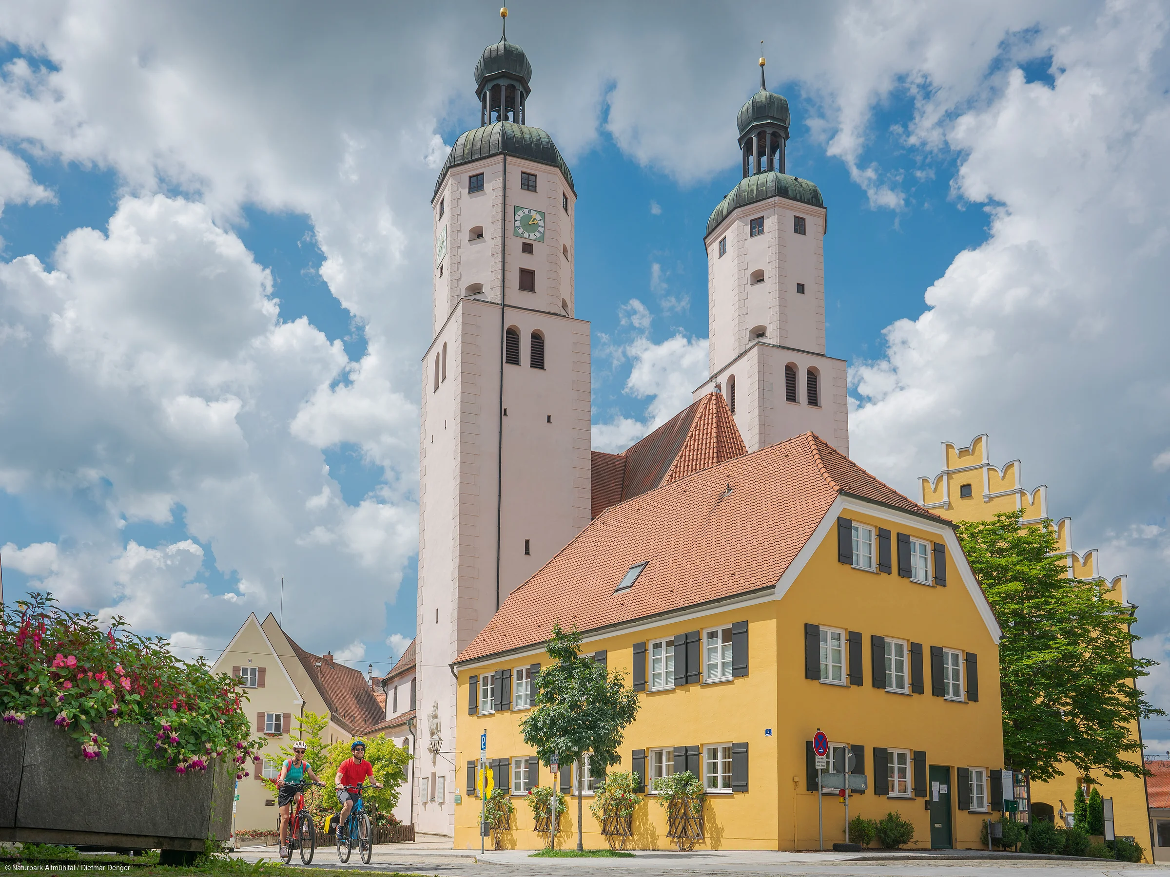 Zwei Radfahrer fahren vor einem gelben Gebäude mit roten Ziegeldächern und einer Kirche mit zwei Türmen im Hintergrund.