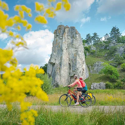 Zwei Radfahrer fahren auf einem Weg vor einem großen Felsen mit Kreuz, umgeben von grüner Landschaft und gelben Blumen.