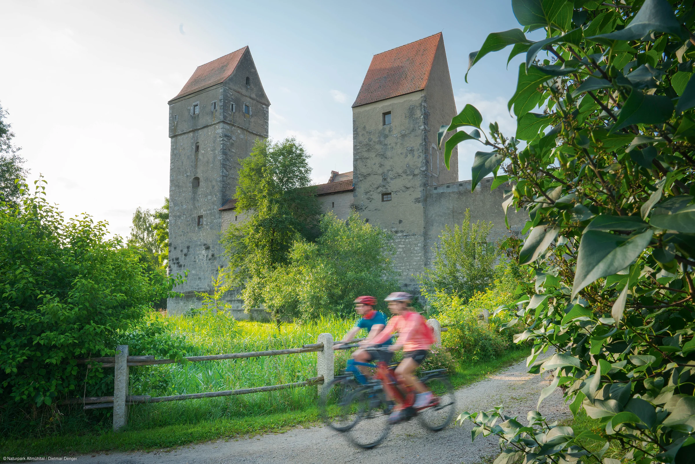Zwei Radfahrer fahren auf einem Weg an einer alten Burg mit zwei Türmen vorbei, umgeben von Bäumen und Sträuchern.