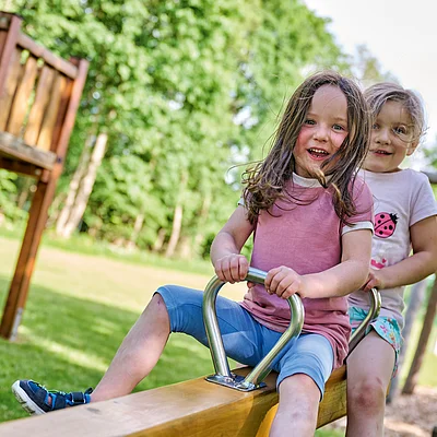 Zwei Mädchen sitzen lachend auf einer Wippe auf einem Spielplatz im Freien bei Sonnenschein.