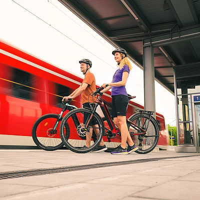Zwei Radfahrer mit Helmen stehen mit ihren Fahrrädern an einem Bahnsteig, im Hintergrund fährt ein roter Zug vorbei.