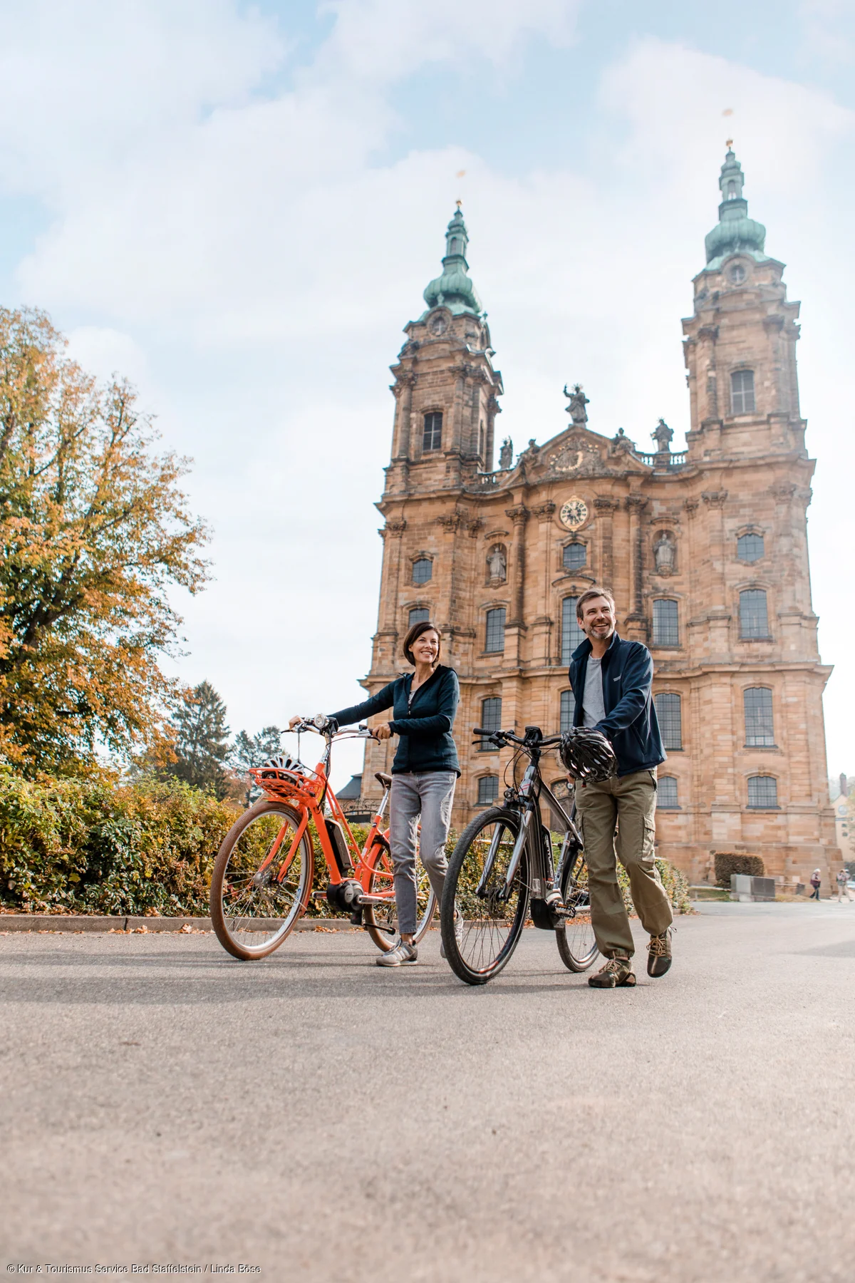 Zwei Personen mit Fahrrädern vor einer großen historischen Kirche an einem sonnigen Tag.