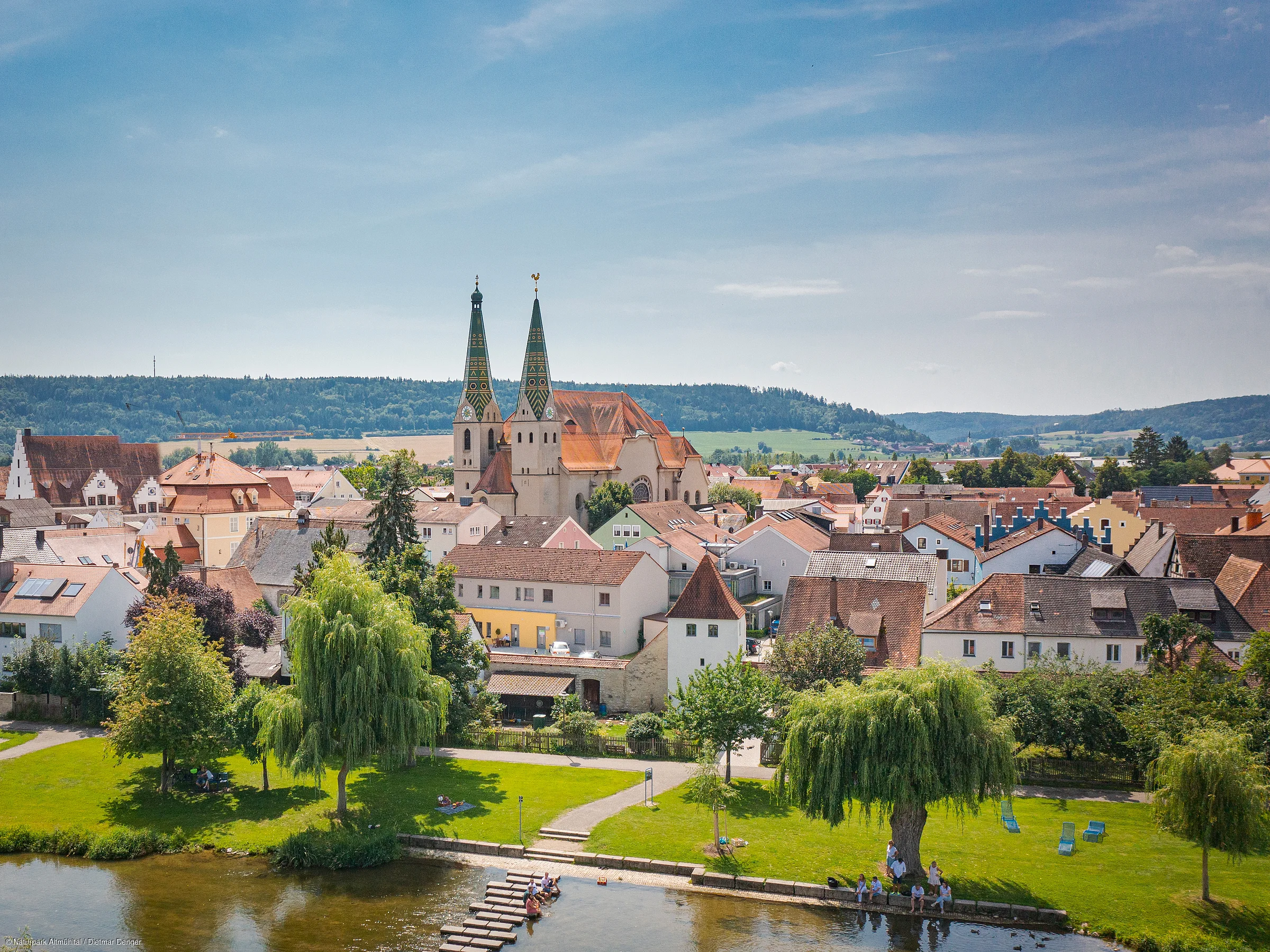 Stadtansicht mit Kirche, Wohnhäusern und Fluss im Vordergrund, umgeben von grüner Landschaft.