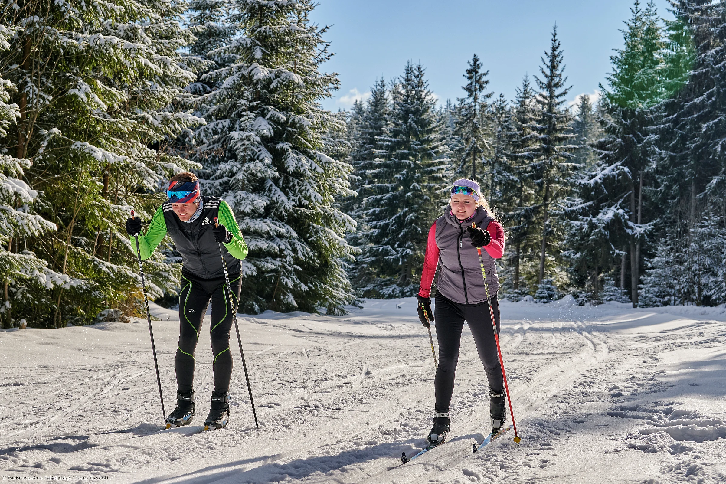 Zwei Personen beim Langlaufen auf verschneiter Waldloipe bei Sonnenschein und blauem Himmel.