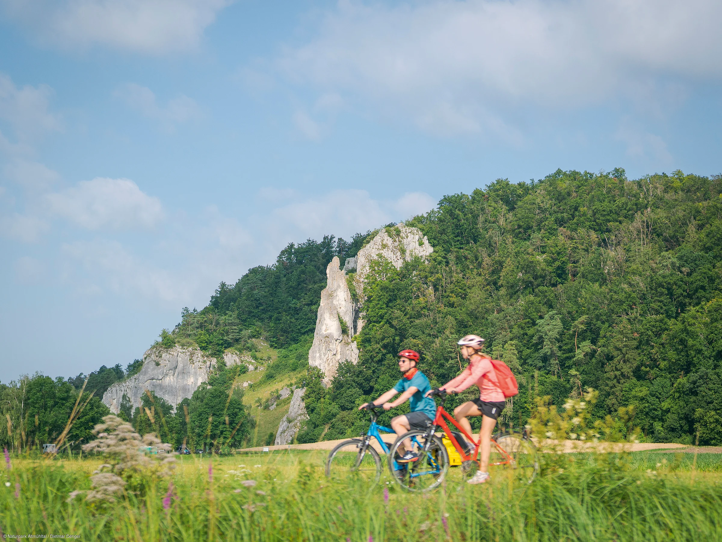 Zwei Personen fahren mit Fahrrädern auf einem Weg vor einer bewaldeten Felslandschaft.