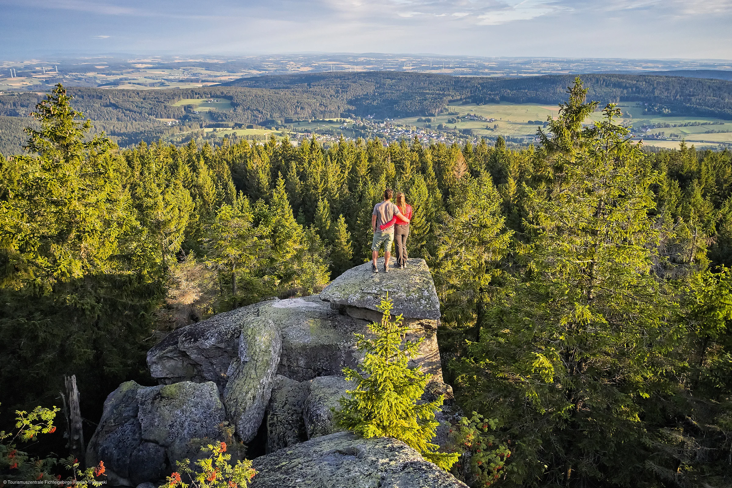 Zwei Personen stehen auf einem Felsen und blicken auf einen Wald und eine Landschaft mit Dörfern und Feldern.