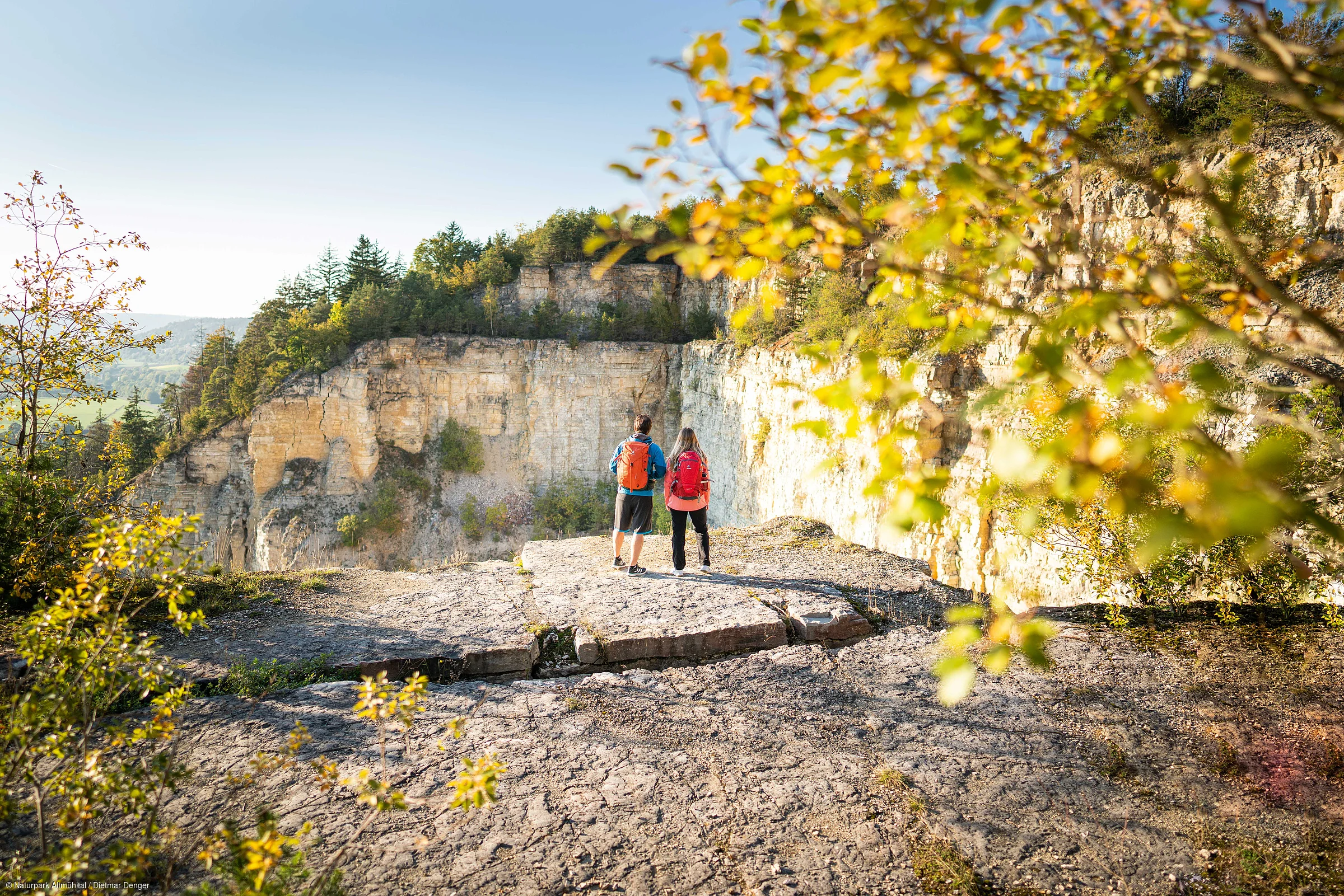 Zwei Personen mit Rucksäcken stehen auf einem Felsvorsprung und blicken auf eine bewaldete Felswand.