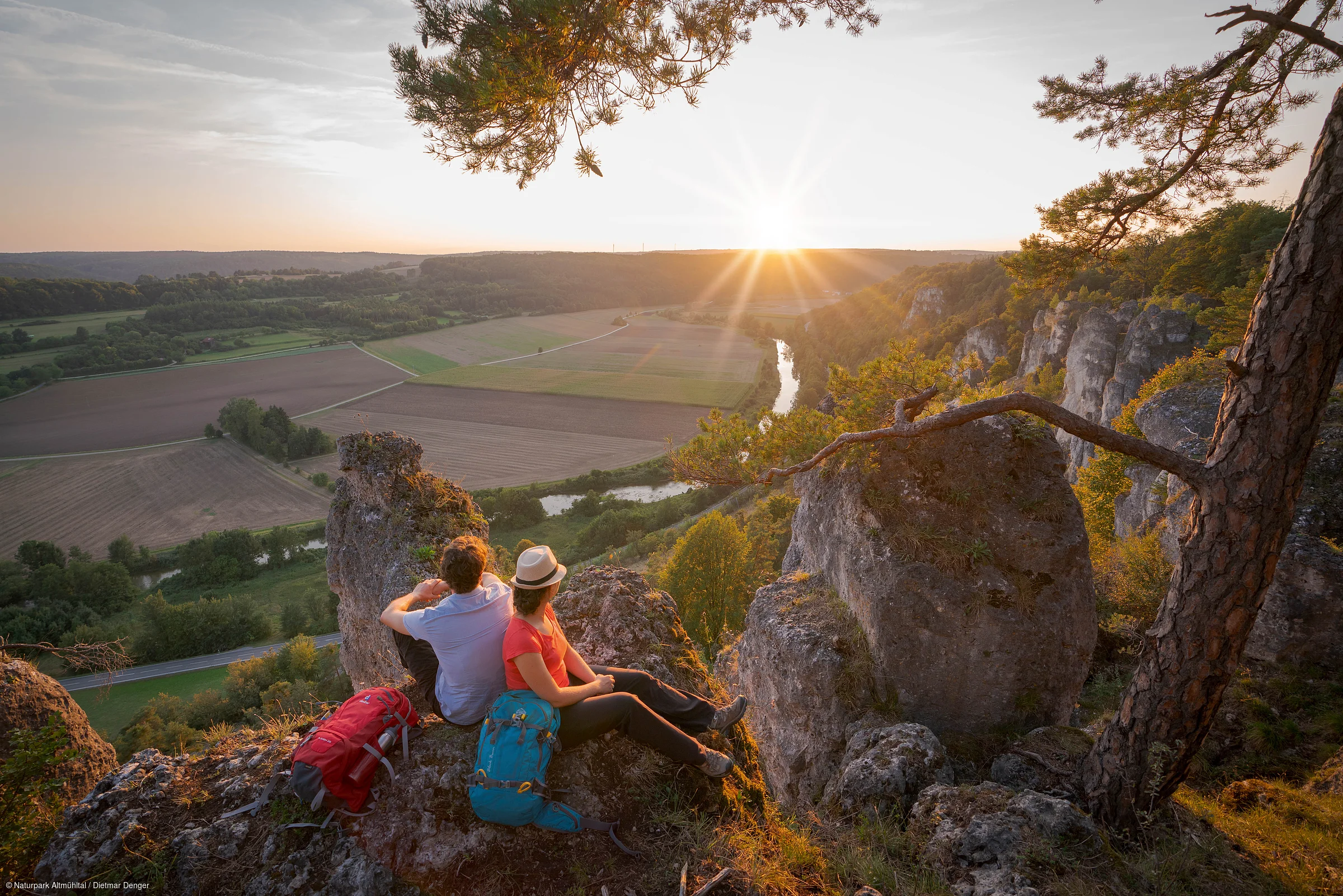 Zwei Wanderer sitzen auf Felsen mit Rucksäcken und blicken auf ein Tal mit Fluss und Sonnenuntergang.