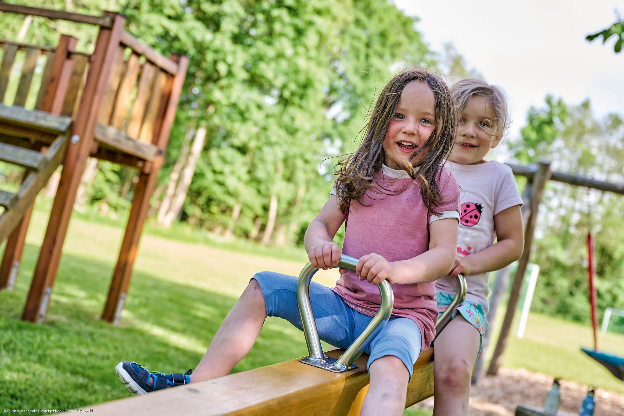 Zwei Mädchen sitzen lachend auf einer Wippe auf einem Spielplatz im Freien bei Sonnenschein.