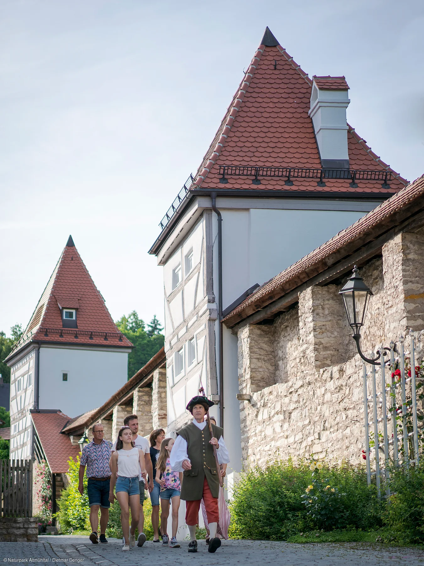 Gruppe mit historisch gekleidetem Stadtführer vor Fachwerkhäusern und Stadtmauer mit Türmen und Laterne.