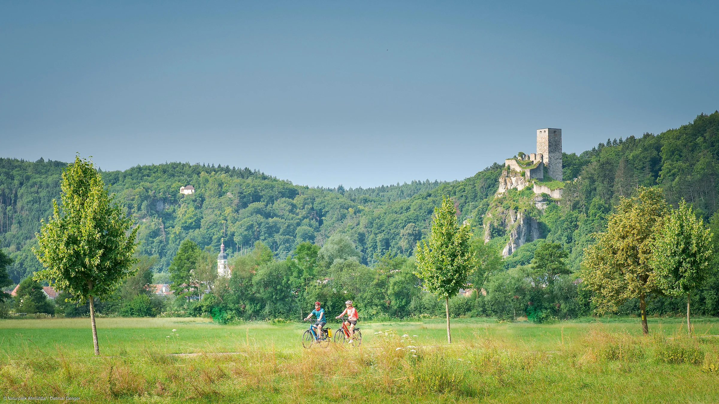 Zwei Personen fahren mit Fahrrädern auf einem Weg durch eine grüne Landschaft mit Bäumen und einer Burgruine im Hintergrund.