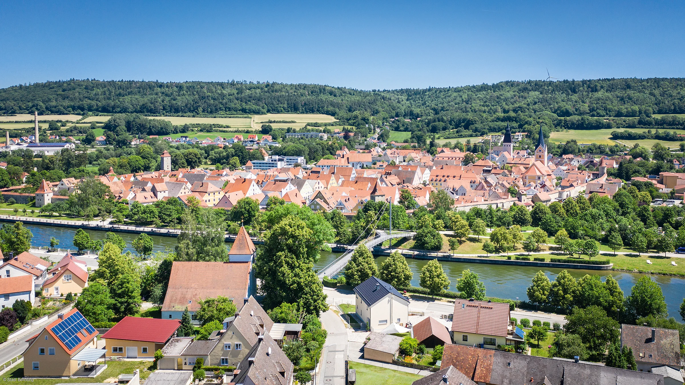 Stadtansicht mit Fluss, Brücke und roten Dächern, umgeben von Bäumen und Hügeln im Hintergrund.