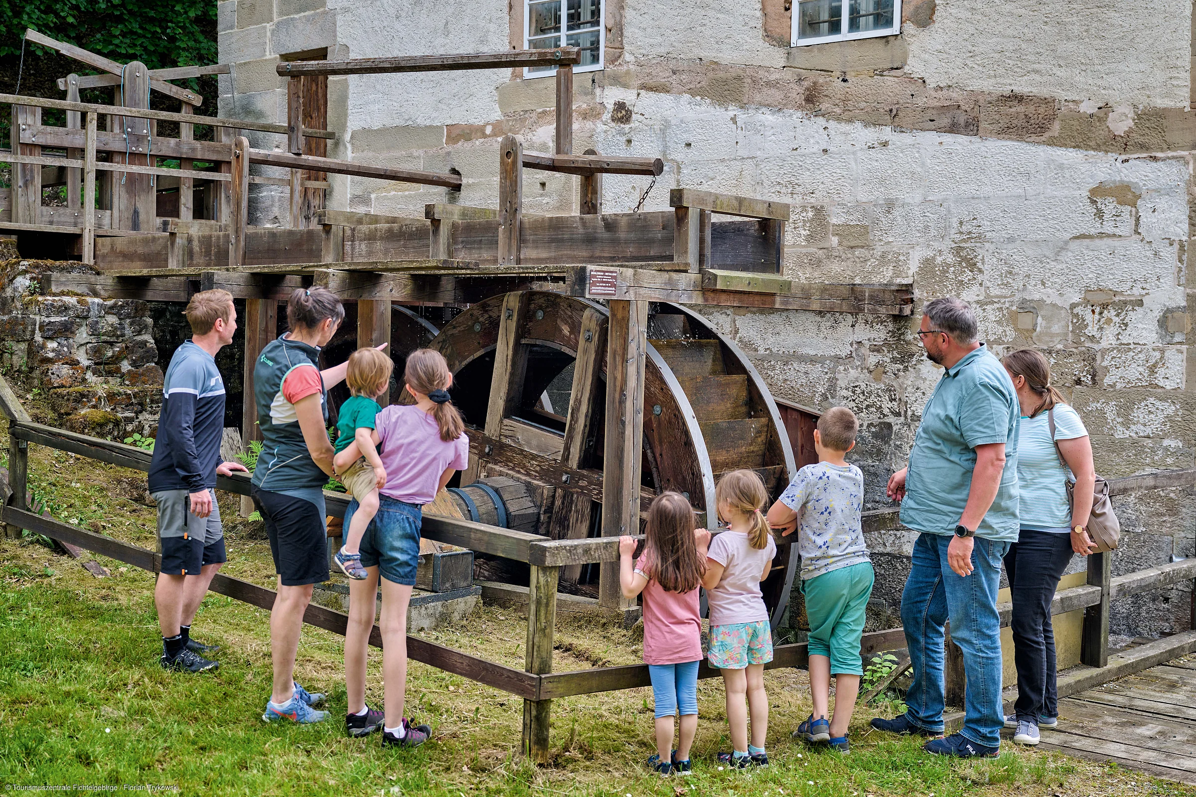 Gruppe von Erwachsenen und Kindern betrachtet historisches Wasserrad an altem Gebäude im Freien.