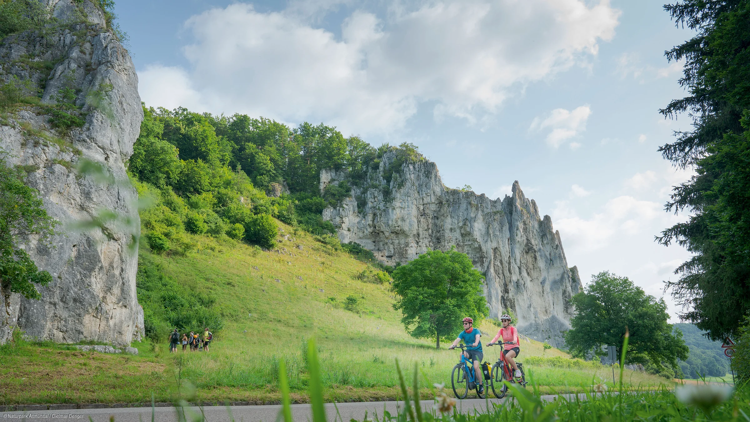 Zwei Radfahrer fahren auf einem Weg vor einer Felsformation und grüner Landschaft.
