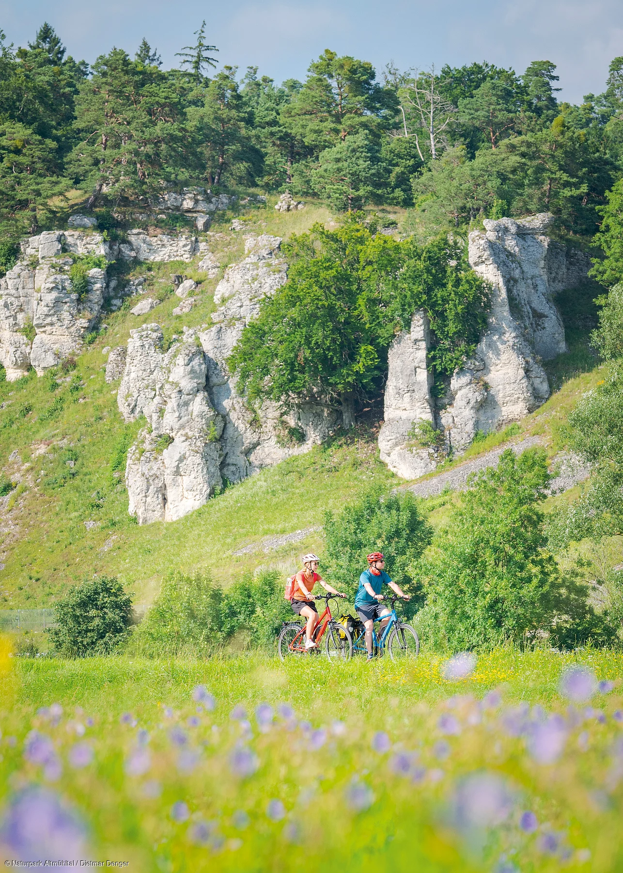 Zwei Personen fahren mit Fahrrädern auf einem Weg vor einer felsigen Landschaft mit Bäumen.