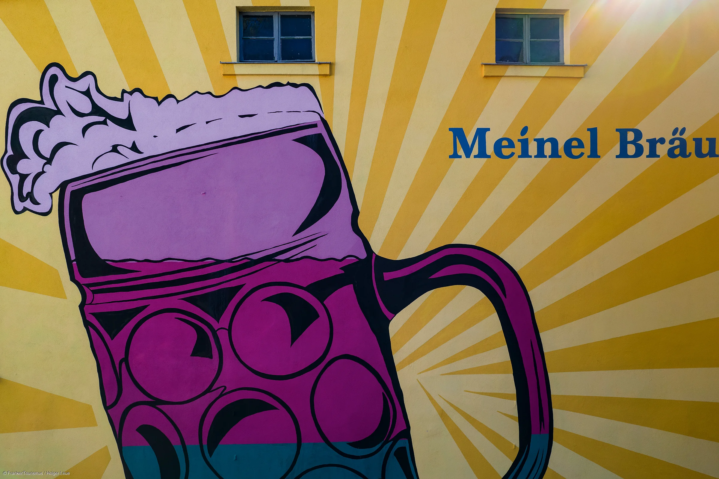 Wandgemälde mit großem lila Bierkrug und Schriftzug „Meinel Bräu“ auf gelb gestreiftem Hintergrund