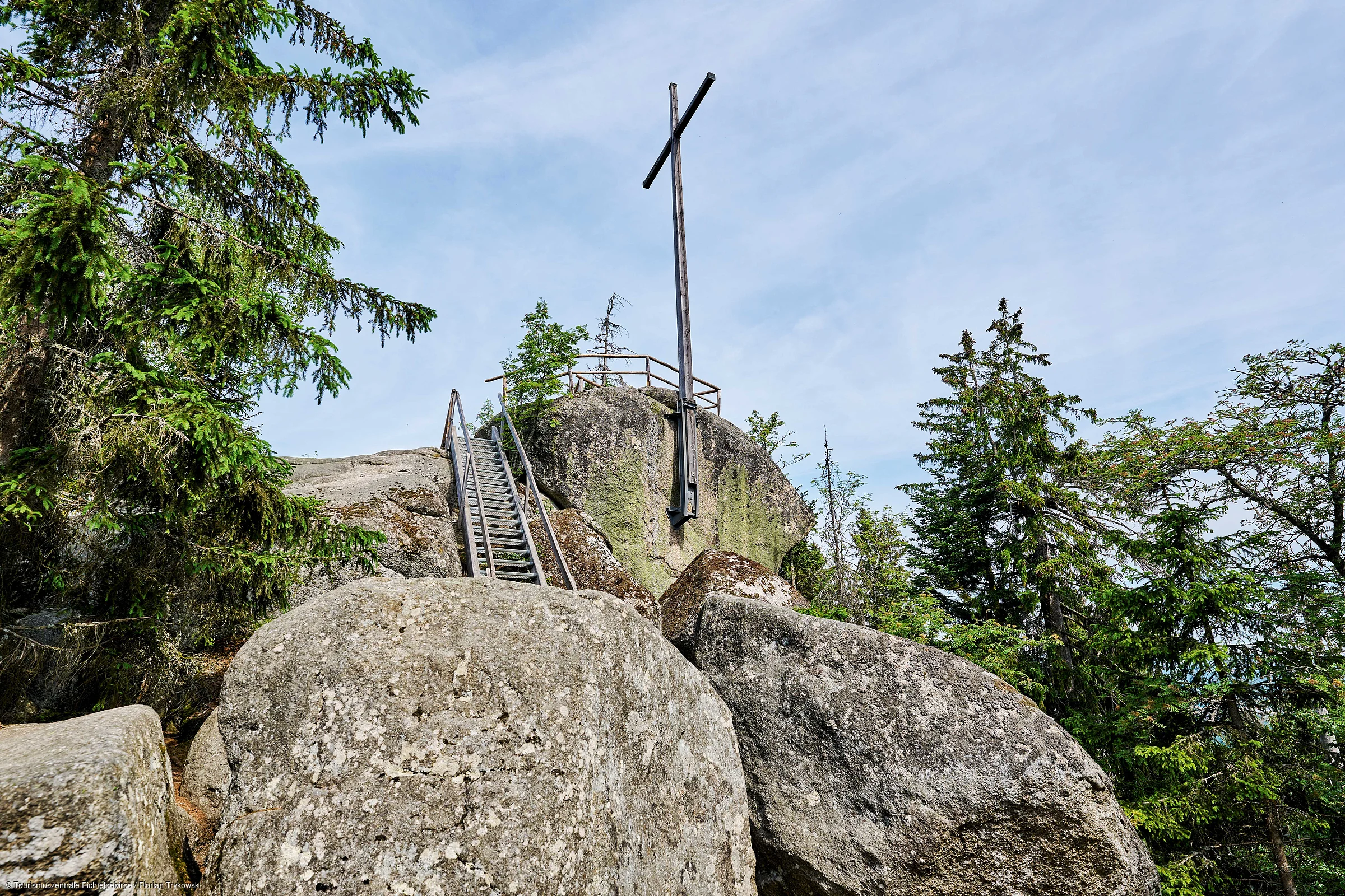 Holzkreuz auf Felsen mit Metalltreppe und umgeben von Nadelbäumen unter blauem Himmel