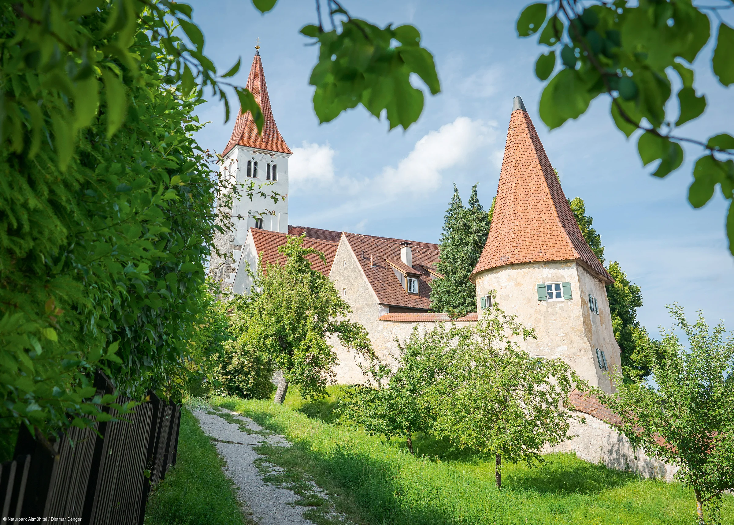 Weg führt an Bäumen vorbei zu historischen Gebäuden mit roten Dächern und Kirchturm unter blauem Himmel
