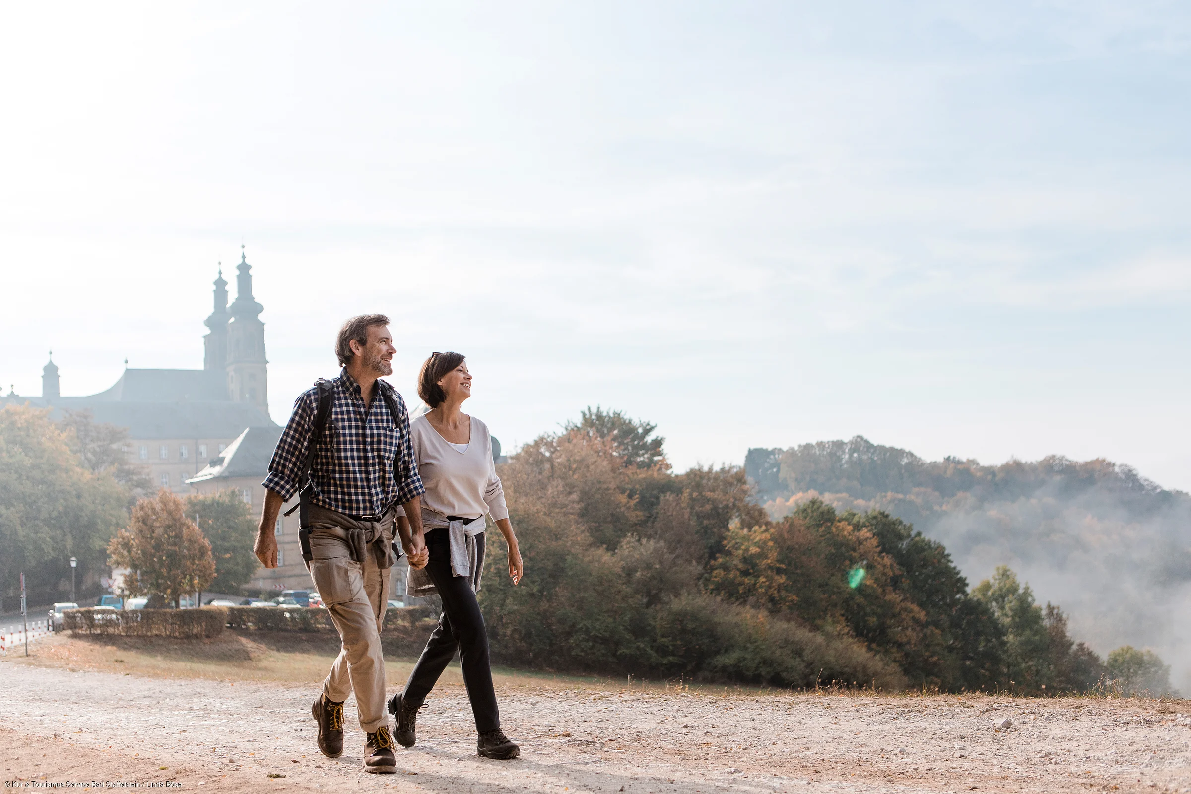 Paar mittleren Alters hält Händchen beim Spaziergang auf einem Weg mit Bäumen und Gebäuden im Hintergrund.