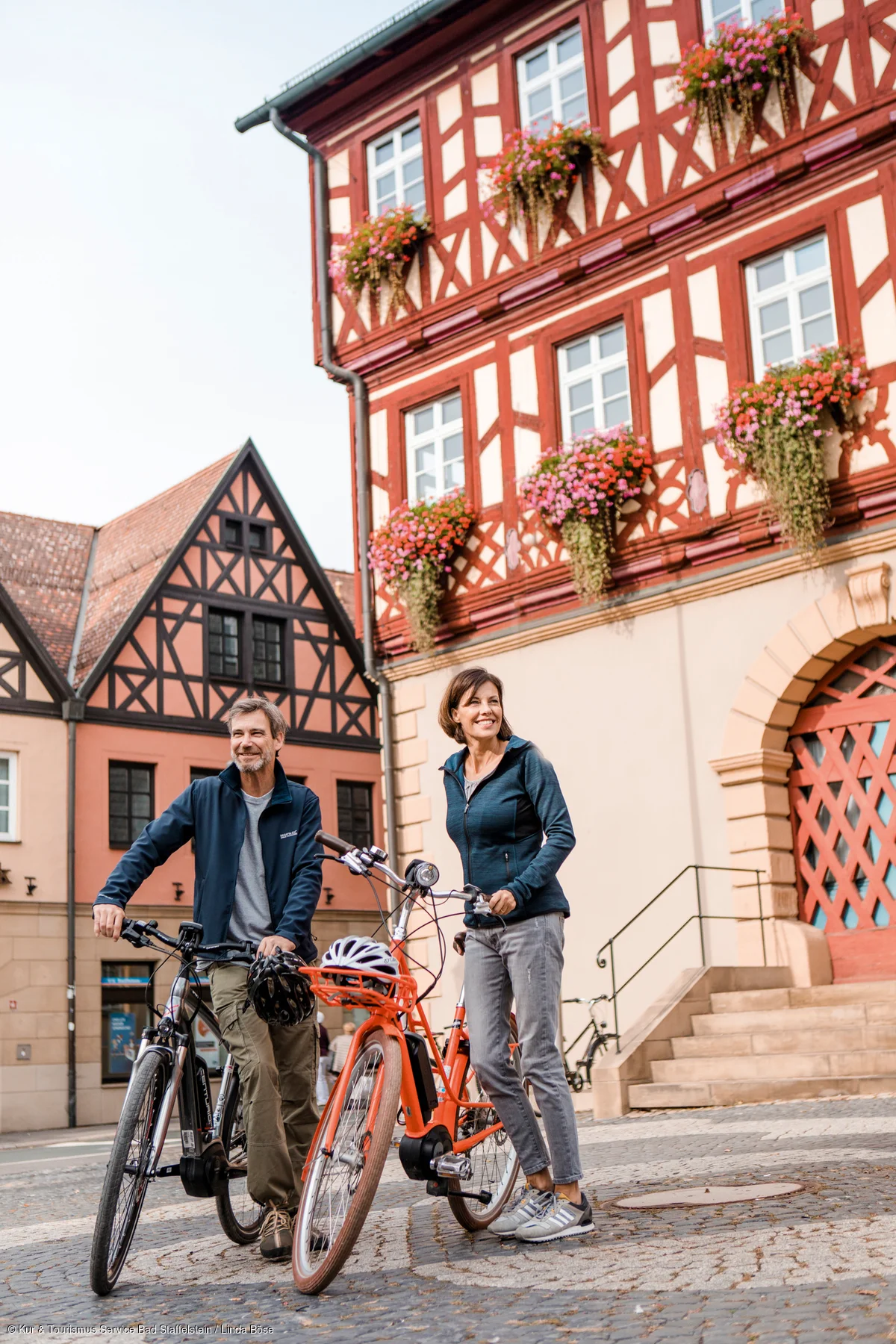 Mann und Frau mit Fahrrädern stehen auf gepflastertem Platz vor Fachwerkhäusern mit Blumenkästen.