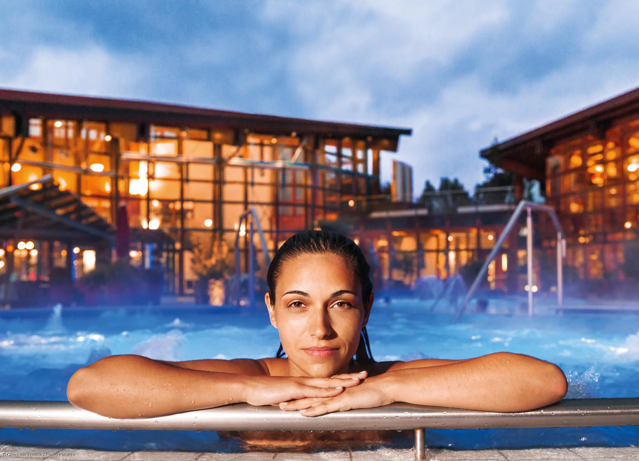 Frau lehnt mit Armen auf Poolrand, im Hintergrund beleuchtete Gebäude bei Abenddämmerung.