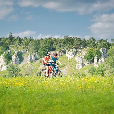 Zwei Personen fahren mit Fahrrädern durch eine grüne Wiese, im Hintergrund Felsen und Bäume.