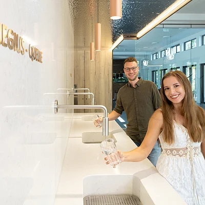 Zwei Personen füllen in modernem Innenraum an Wandwasserhähnen Gläser mit Wasser auf.