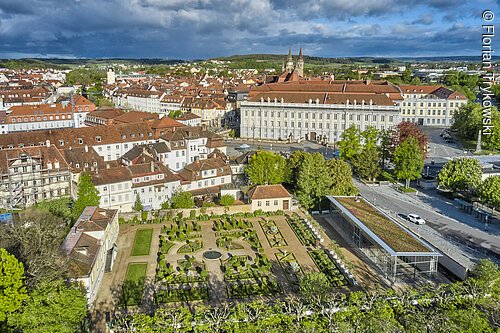 Luftaufnahme eines barocken Gartens mit angrenzenden historischen Gebäuden und Stadt im Hintergrund bei bewölktem Himmel.
