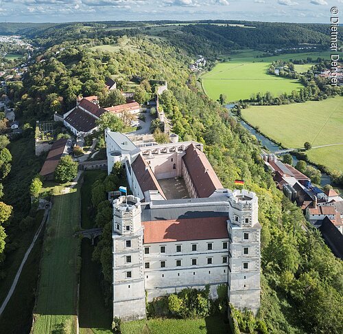 Luftaufnahme einer Burg mit zwei Türmen, umgeben von Wald und Feldern, bei klarem Himmel.