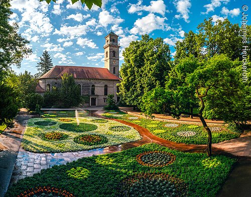 Park mit bunten Blumenbeeten, einem Baum und einem Gebäude mit Uhrenturm bei blauem Himmel und Wolken.