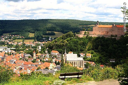 Blick auf Stadt mit Kirche, Burg auf Hügel und Wald im Hintergrund, davor eine leere Holzbank auf Wiese.