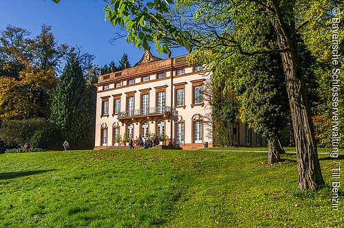 Schloss Schönbusch (Aschaffenburg, Spessart-Mainland) Klassisches Gebäude mit großen Fenstern auf einer grünen Wiese, umgeben von Bäumen bei klarem Himmel.