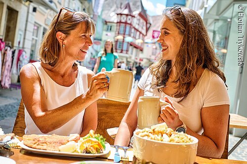 Fränkische Köstlichkeiten (Ansbach, Romantisches Franken) Zwei Frauen stoßen mit Bierkrügen an, sitzen draußen an einem Tisch mit Essen in einer belebten Fußgängerzone.
