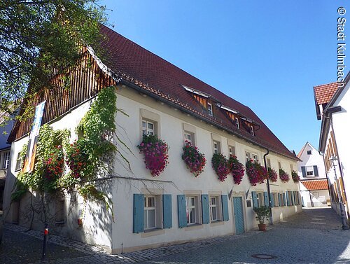 Weißes zweistöckiges Haus mit blaugrünen Fensterläden und Blumenkästen an einer gepflasterten Straße bei klarem Himmel