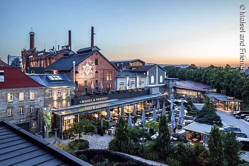 Außenansicht eines Brauereigebäudes mit Terrasse und Garten bei Abendlicht, umgeben von Bäumen und Parkplätzen.