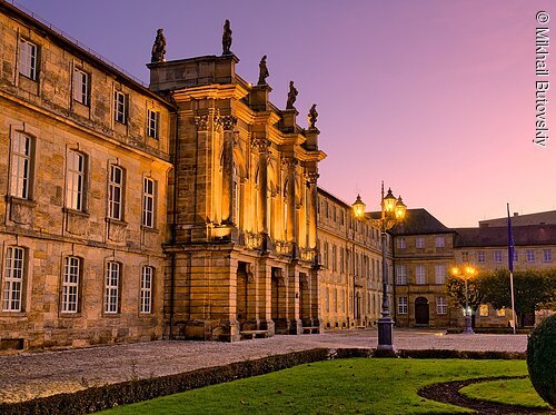 Historisches Gebäude mit Säulen und Statuen bei Sonnenuntergang, beleuchtete Straßenlaternen und gepflegter Garten.
