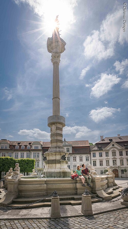 Steinerner Brunnen mit Säule und Figuren, zwei Personen sitzen am Rand, historische Gebäude im Hintergrund.