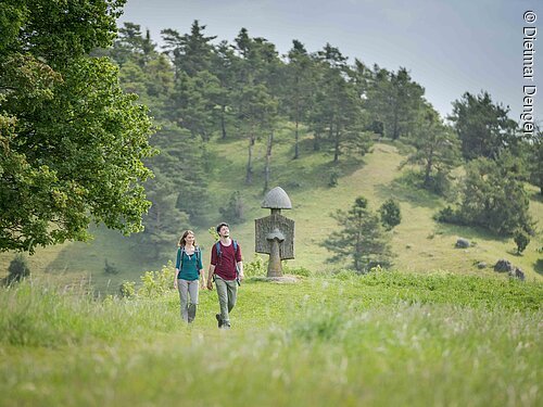 Paar mit Rucksäcken wandert auf einem Grasweg mit einem steinernen Wegweiser und bewaldetem Hügel im Hintergrund.