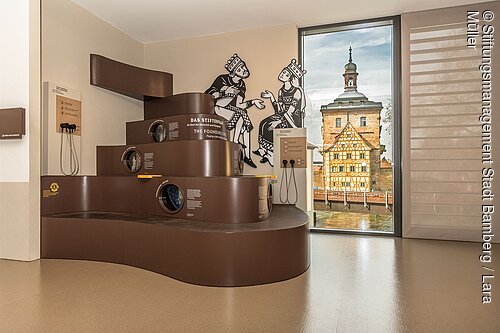 Welterbe-Besuchszentrum (Bamberg, Steigerwald) Museumsausstellung mit braunem Stufenmodell, Hörstationen und Fenster mit Blick auf Fachwerkhaus und Turm
