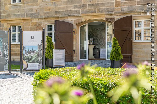 Infozentrum St. Michael (Bamberg, Steigerwald) Eingang eines Sandsteingebäudes mit offenem Tor, Schildern und bepflanzten Töpfen davor, unscharfe Blumen im Vordergrund.