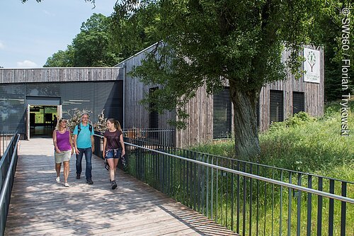Drei Personen gehen auf einem Holzsteg zu einem Gebäude mit Holzfassade und Schild „Steigerwald Zentrum“.
