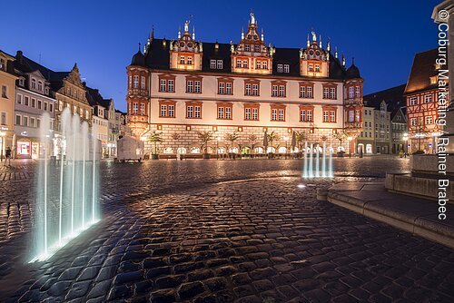Beleuchteter Marktplatz mit historischen Gebäuden und zwei beleuchteten Wasserspielen bei Abenddämmerung.