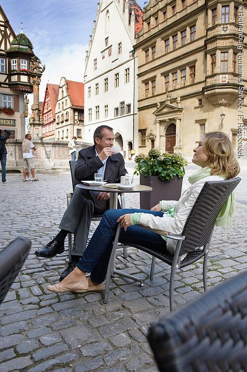 Zwei Personen sitzen an einem Tisch auf gepflastertem Platz vor historischen Fachwerkhäusern und trinken Kaffee.