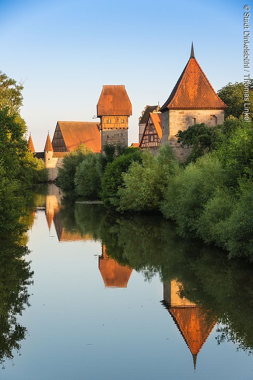 Historische Fachwerkhäuser und Türme spiegeln sich in einem ruhigen Fluss, umgeben von grünen Bäumen.