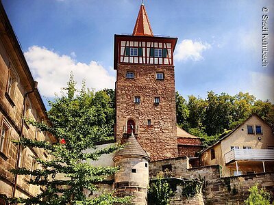 Steinerner Turm mit Fachwerk und rotem Dach vor blauem Himmel und Bäumen im Hintergrund